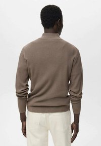 Suéter marrón acanalado con un cuello alto, mangas largas y dobladillo ceñido. Usado sobre pantalones de color crema, mostrando un patrón de tejido texturizado.