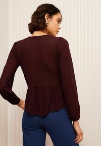 Burgundijska bluzica dugih rukava s peplum rubom i teksturiranim materijalom. Ima valoviti rub i opušteni kroj. Nosila se uz plave traperice.