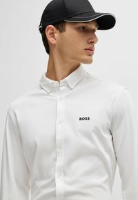White long-sleeve shirt with a button-down collar, featuring a black logo "BOSS" on the chest. Paired with a black cap with a white accent.