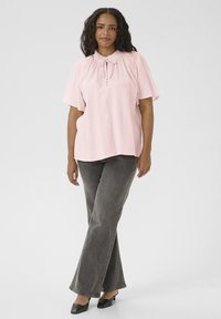 Blouse rose à manches courtes avec col volanté et détail à nouer, associée à un jean évasé gris et des chaussures à glisser noires. Texture lisse dans l'ensemble.