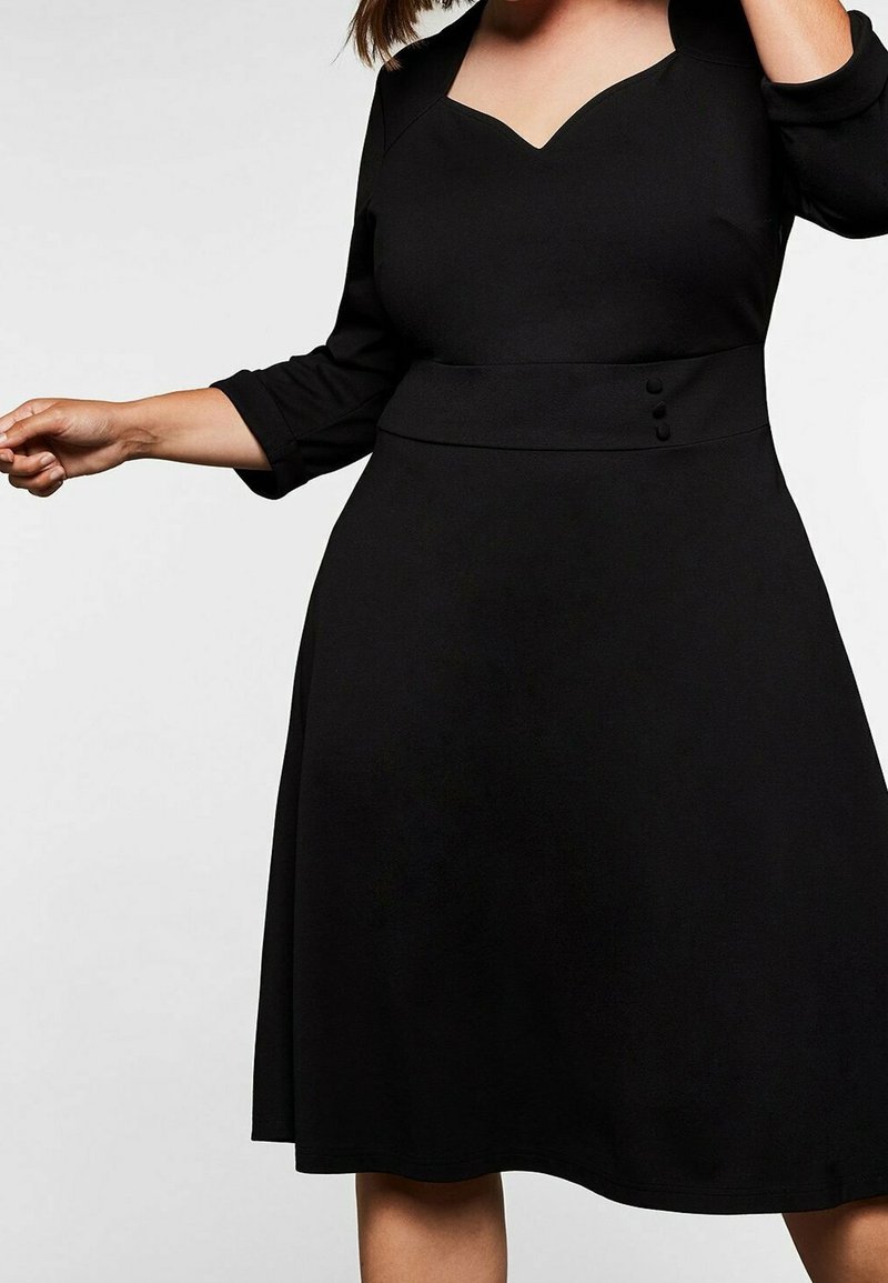 Femme portant une robe noire trapèze avec des manches trois-quarts, un décolleté en cœur et trois boutons décoratifs à la taille.