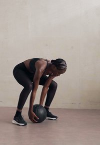 Persoon in zwarte sportkleding, gebogen om een zwarte medicijnbal op te tillen. De achtergrond is eenvoudig, met lichte grijze wand- en vloerteksten.