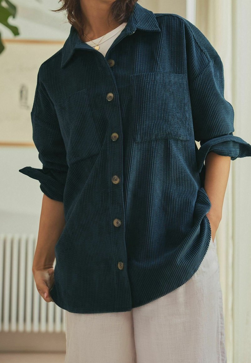 Personne portant une chemise en velours côtelé bleu foncé boutonnée avec les manches retroussées sur un haut blanc et un pantalon beige clair, debout à l'intérieur.