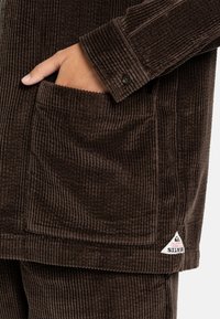 Brown corduroy jacket featuring vertical ribbed texture, a buttoned cuff, and a front pocket with a logo patch at the hem.