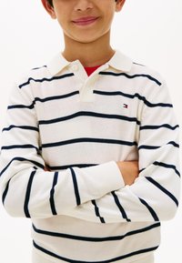 Αγόρι που φοράει λευκό πόλο με μακριά μανίκια και ναυτικές οριζόντιες ρίγες, στέκεται με σταυρωμένα τα χέρια και ελαφρύ χαμόγελο.