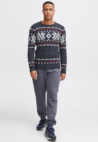 Gebreide trui in donkergrijs met een feestelijk wit en bordeauxrood sneeuwvlokpatroon; gedragen met grijze joggingbroek en blauwe sneakers.