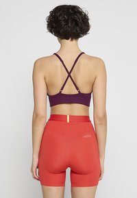 Sporta krūšturis tumši violetā krāsā ar racerback dizainu, ko papildina pieguļoši sarkani šorti ar dzeltenu akcentu jostasvietā.