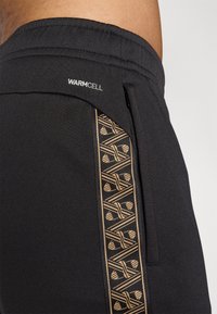 Melnas sporta bikses ar teksturētu audumu, sānu kabatu un Warmcell simbolu tuvu jostasvietai. Zeltainas ģeometriskas formas raksts sānos.