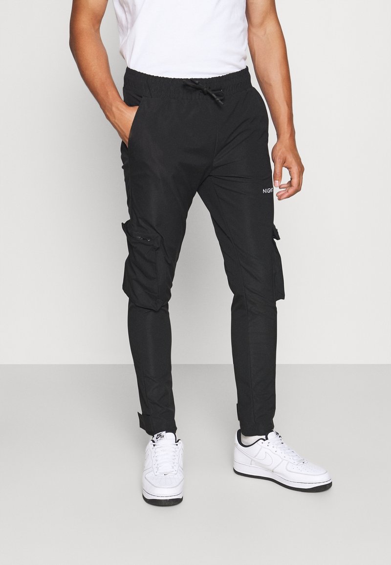 Homme portant un pantalon cargo noir avec taille élastique et des baskets blanches, debout devant un fond clair uni.