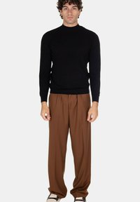 Uomo in piedi, con un maglione nero a maniche lunghe, pantaloni larghi marroni e sneakers bianco e nere, sullo sfondo bianco.