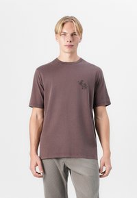T-shirt à manches courtes, col rond en coton marron neutre, avec un petit graphique noir sur la poitrine gauche. Finition texturée.