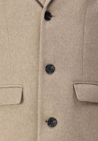 Beige Wollmantel mit strukturiertem Finish, drei schwarzen Knöpfen und zwei Fronttaschen mit Klappen. Geeignet für kühleres Wetter.