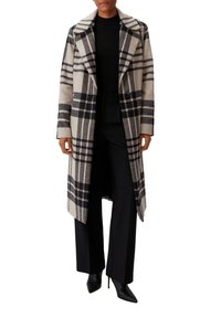 Beige-schwarz karierter Wollmischungsmantel mit breitem Revers, doppelter Knopfleiste und lockerer Passform, kombiniert mit schwarzen Hosen und Absätzen.