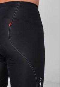 Leggings de sport noirs avec une texture lisse, présentant des coutures blanches, une taille haute et un petit logo rouge près de la hanche.