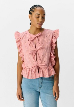 Blouse rose brodée avec des épaules volantes, des nœuds à l'avant et un ourlet en couches. Associée à un jean en denim bleu clair.