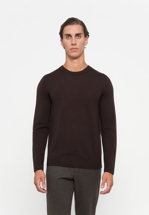 Man wearing a dark brown long-sleeve crewneck sweater and gray pants, standing against a plain white background.