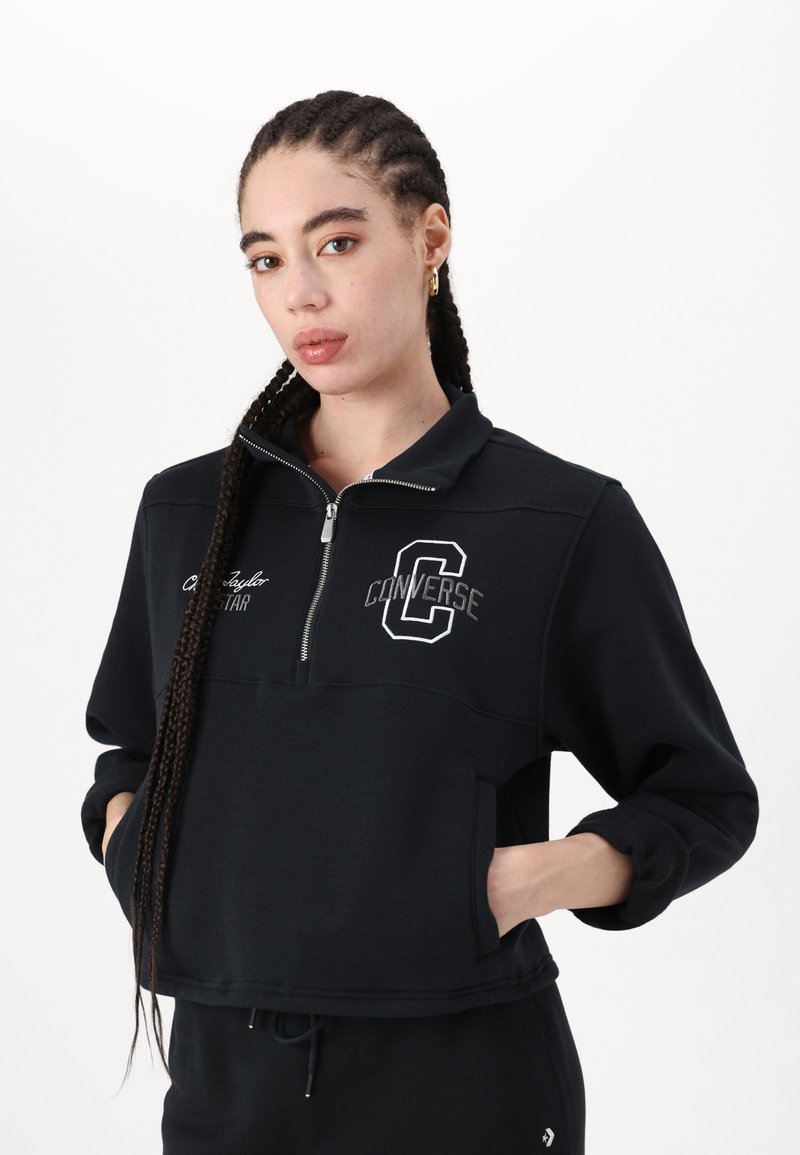 Sweatshirt noir court avec un col zippé, logo "Converse" brodé et poches kangourou ; texture douce et design à coupe décontractée.