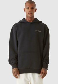 Hanorac negru din amestec de bumbac. Prezintă o buzunar frontal, manșete ribbate și un mic logo alb pe pieptul stâng. Design simplu, croială lejeră.