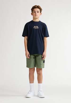 T-shirt en coton bleu marine avec texte crème, associé à un short cargo vert olive, des baskets blanches et des chaussettes blanches mi-mollet.