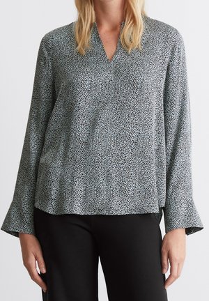 Vrouw draagt een lichtblauw blouse met lange mouwen met kleine zwarte luipaardvlekken en zwarte broek, staand tegen een effen achtergrond.