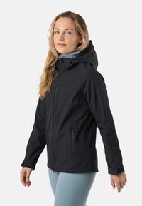 Femme aux longs cheveux blonds portant une veste à capuche noire et un pantalon bleu clair, debout de profil devant un fond uni.
