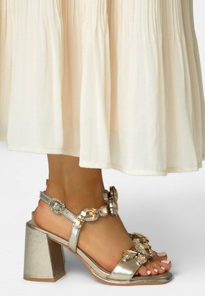 Pied portant une sandale à talon bloc argenté ornée de grandes pierres précieuses transparentes, associée à une jupe fluide couleur crème.