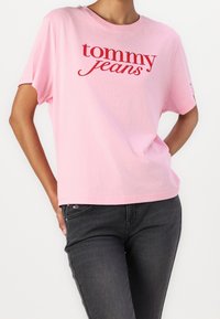 Camiseta de algodón de color rosa con mangas cortas, que presenta un logo rojo "tommy jeans" en el pecho. Combinada con unos jeans gris oscuro.