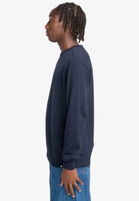 Dunkelblaues Crewneck-Sweatshirt aus weichem Stoff. Mit gerippten Bündchen und Saum, taillierter Silhouette und minimalen Nähdetails. Kombiniert mit hellblauen Jeans.
