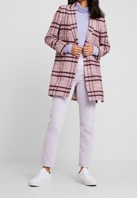 Kariertes langes Sakko in Weinrot und Creme, mit Reverskragen, kombiniert mit einem hellvioletten Oberteil und hellvioletten Jeans, abgerundet mit weißen Sneakern.