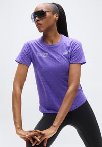 Μωβ αθλητική μπλούζα με κοντά μανίκια, στρογγυλή λαιμόκοψη και ανακλαστικές λεπτομέρειες λογότυπου. Φορεμένη με μαύρο εφαρμοστό κολάν και μεγάλα γυαλιά ηλίου.