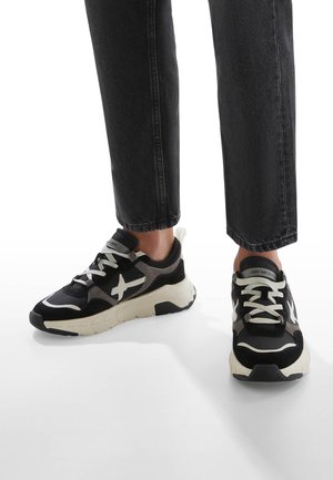 Schwarz-weiße Sport Sneakers mit strukturiertem Gewebe, weißen Schnürsenkeln und klobigen beigen Sohlen. Mit einem Logo-Akzent an der Seite.