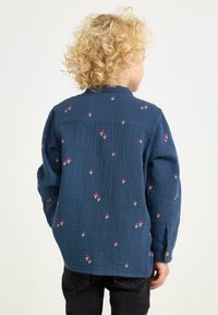 Blå bomullsskjorta med knappar som har röd svampbroderi, texturerad tyg, långa ärmar och rundad nederkant.