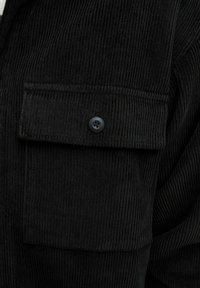 Tissu en velours côtelé noir avec une poche à rabat fermée par un seul bouton noir sur un vêtement.