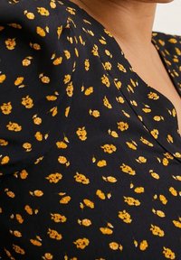 Tissu noir à imprimé floral jaune avec un design à col en V et des manches bouffantes. Texture lisse avec de petits accents floraux dispersés.