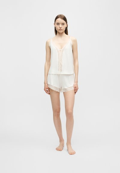 Jeune femme debout, pieds nus, portant un ensemble pyjama blanc orné de dentelle, constitué d'un caraco et d'un short, sur un fond clair uni.