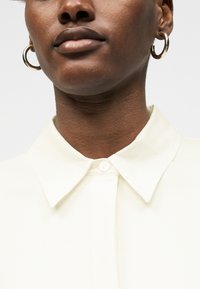 Κοντινό πλάνο ενός ατόμου που φοράει χρυσούς κρίκους στα αυτιά και μια κρεμ πουκαμισοειδή μπλούζα με κουμπωμένο το πάνω κουμπί.