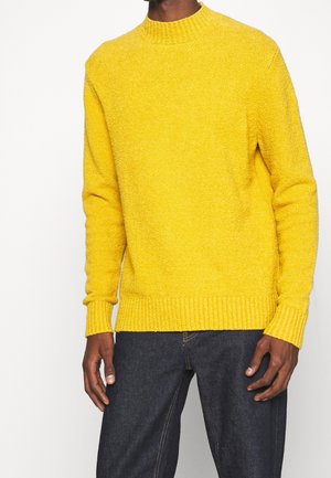 Gelber Strickpullover mit hohem Kragen, gerippten Bündchen und Taille. Weicher, strukturierter Stoff, kombiniert mit dunklen Jeans.