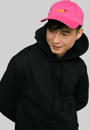 Różowa czapka bejsbolowa z haftowanym wzorem jamnika, noszona na czarnym hoodie. Czapka ma zakrzywiony daszek i otwory wentylacyjne.