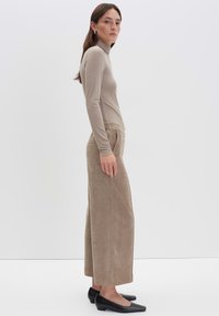 Femme en pull col roulé beige et pantalon en velours côtelé large, portant des chaussures noires à talon bas, se tenant de profil contre un fond blanc uni.
