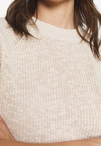 Mujer vestida con un suéter de punto color crema con cuello acanalado y un patrón texturizado, con cabello castaño oscuro hasta los hombros.