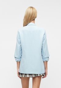 Svetlomodrý blazer so štruktúrovaným dizajnom, s vyvalenými rukávmi a jedným rozparkom. Nosený cez krátku, károvanú sukňu. Hladká textúra látky.