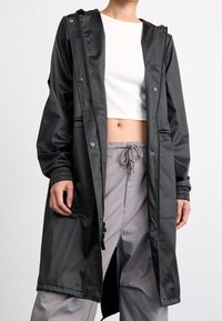 Schwarzer, wasserdichter langer Mantel mit Kapuze, Druckknöpfen, elastischen Bündchen und zwei Vorderentaschen, kombiniert mit hellgrauer Hose.