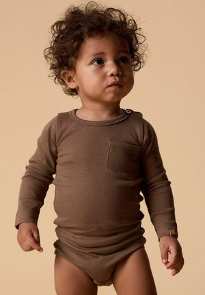 Tout-petit aux cheveux bouclés portant un body côtelé marron à manches longues, regardant légèrement vers la droite sur un fond beige.