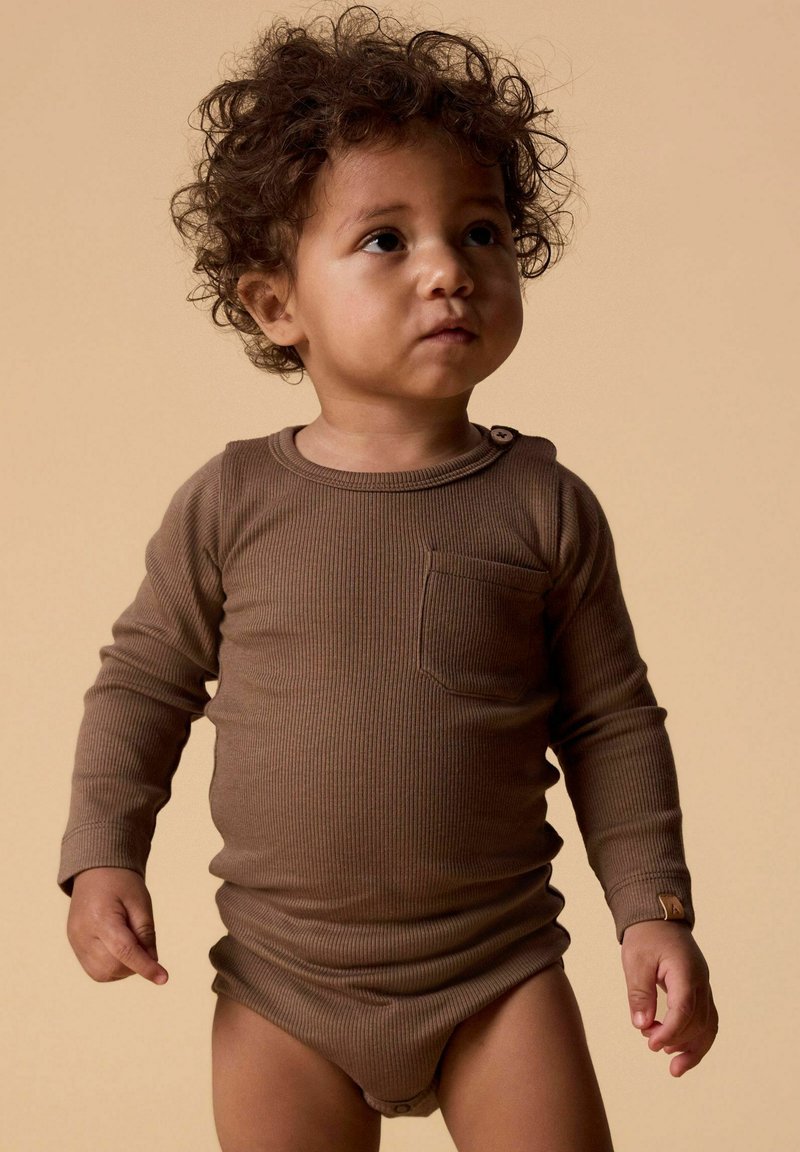 Tout-petit aux cheveux bouclés portant un body côtelé marron à manches longues, regardant légèrement vers la droite sur un fond beige.