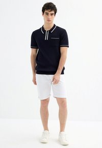 Polo de marino acanalado con acentos blancos y bolsillo, combinado con pantalones cortos blancos remangados y zapatillas blancas. Diseño simple y moderno.
