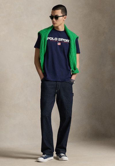 Μπλε φανελάκι με το κείμενο "POLO SPORT", πράσινο πλεκτό γιλέκο, σκούρα τζιν και μπλε αθλητικά παπούτσια με λευκές σόλες. Αξεσουάρ περιλαμβάνουν γυαλιά ηλίου.