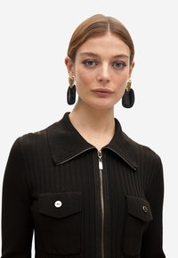 Vestido de punto acanalado negro con cierre de cremallera, cuenta con un collar y dos bolsillos delanteros. Accesorio con grandes pendientes negros y dorados.
