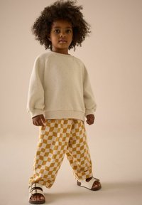 Helles cremefarbenes Sweatshirt mit strukturiertem Stoff und Raglanärmeln, kombiniert mit gelb-weiß karierten Hosen. Weiße Sandalen mit Schnallen.