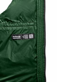 Groene gewatteerde jas met een ritszak met een zwart etiket en QR-code. Soepele textuur, lichtgewicht ontwerp en doorgestikte naden.