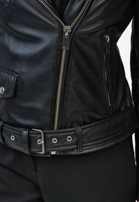 Zwarte leren jack met zilveren ritsen, een ceintuur in de taille en twee ritszakken. Gladde textuur met een aansluitend design.